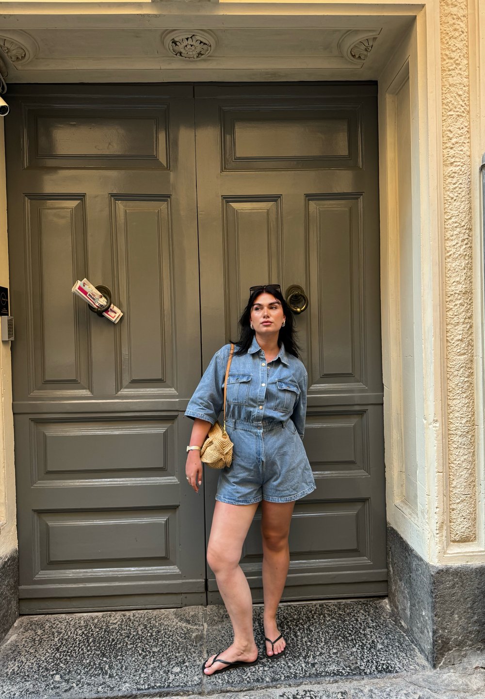 Τζιν ολόσωμο φόρεμα με κοντά μανίκια, ανοιχτό μπλε χρώμα, σφιχτή μέση και δύο τσέπες στο στήθος. Συνδυάζεται με μια διχτυωτή τσάντα και σαγιονάρες. Φόντο με σκούρα ξύλινη πόρτα.