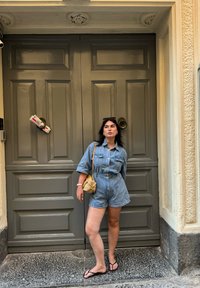 Τζιν ολόσωμο φόρεμα με κοντά μανίκια, ανοιχτό μπλε χρώμα, σφιχτή μέση και δύο τσέπες στο στήθος. Συνδυάζεται με μια διχτυωτή τσάντα και σαγιονάρες. Φόντο με σκούρα ξύλινη πόρτα.