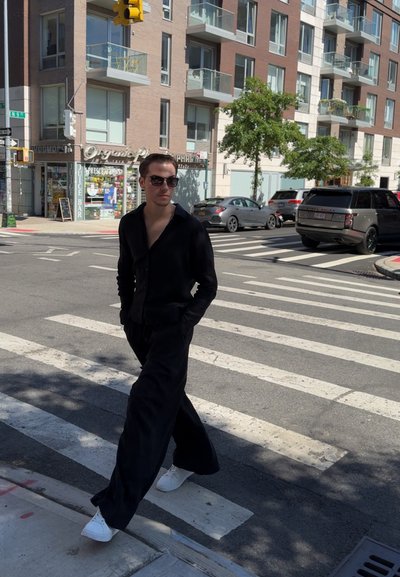 Camisa y pantalones negros, zapatillas blancas, caminando por un paso de peatones. Fondo urbano con un bar de zumos y coches aparcados. Día claro y soleado.