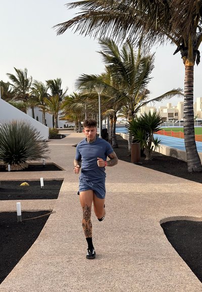 Un hombre tatuado con ropa deportiva azul corre por un camino bordeado de palmeras cerca de una pista de atletismo azul y edificios blancos.