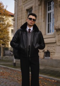 Czarna skórzana kurtka bomberka z futrzanym kołnierzem, szary sweter i czarne spodnie. Stylowe okulary przeciwsłoneczne dopełniają nowoczesny wygląd stroju.