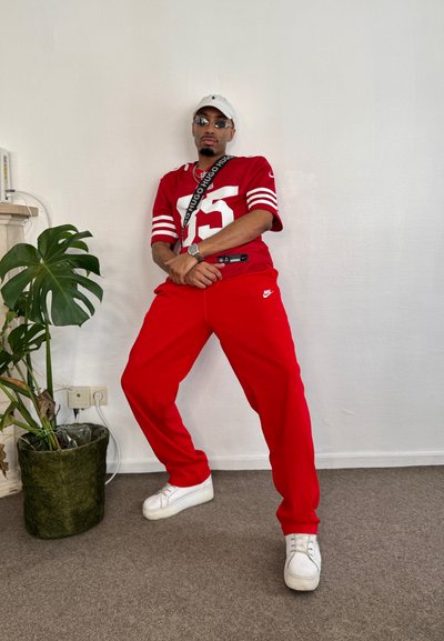 Jersey atlético rojo con rayas blancas y el número 25, combinado con pantalones de chándal rojos y zapatillas blancas. Los accesorios incluyen un bolso cruzado negro.