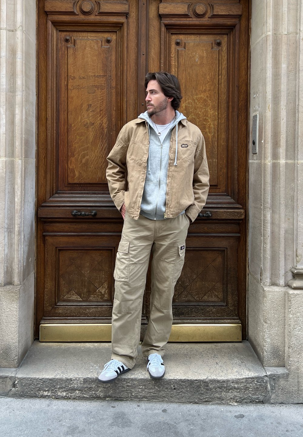 Καφέ καμβά ζακέτα πάνω από γκρι hoodie, μπεζ cargo παντελόνι και λευκά αθλητικά παπούτσια με μαύρες ρίγες, στέκεται μπροστά σε μια ξύλινη πόρτα.