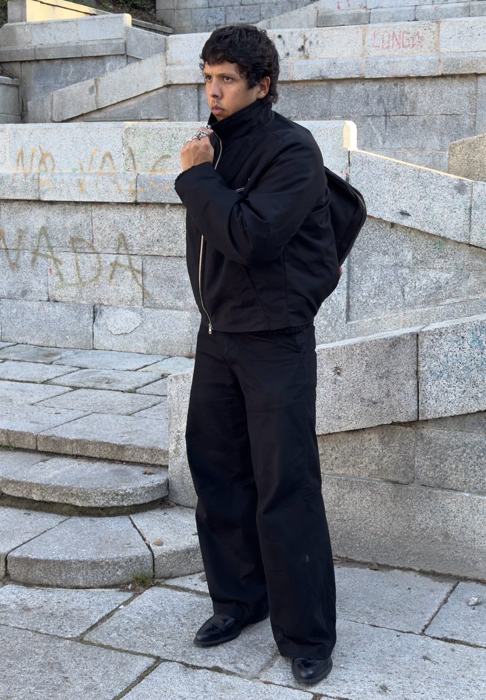 Black zip-up jacket with a high collar, paired with loose black pants and black shoes, standing on stone steps with graffiti in the background.
