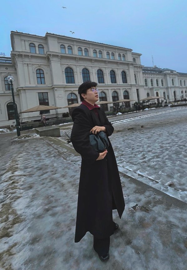 Giovane con cappotto nero che tiene una borsa, in piedi su un marciapiede ghiacciato vicino a un grande edificio classico con finestre ad arco e ombrelloni all'aperto.