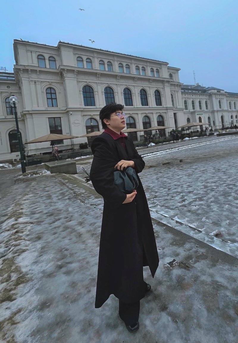 Jeune personne en manteau noir tenant un sac, debout sur un trottoir glacé près d'un grand bâtiment classique avec des fenêtres arquées et des parasols extérieurs.