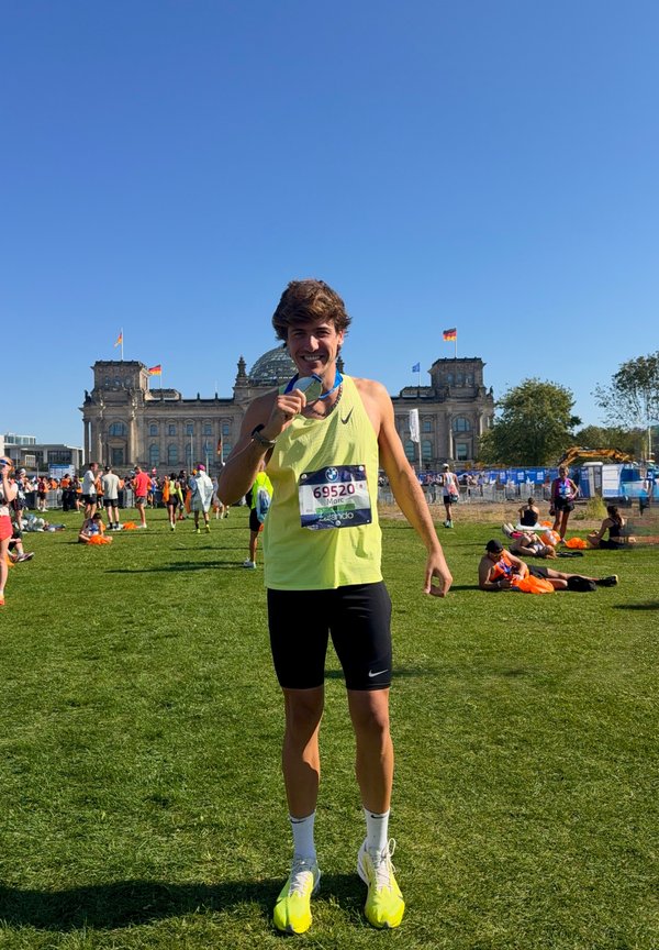 Joven corredor masculino con camiseta sin mangas amarilla y pantalones cortos negros muerde una medalla frente al edificio del Reichstag después de una carrera en un día soleado.