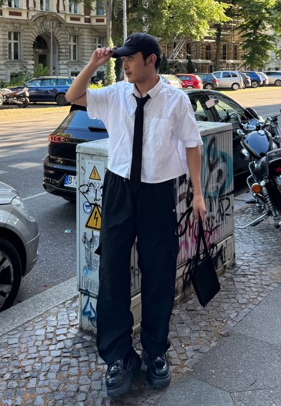 Camisa blanca de manga corta, corbata negra, pantalones negros holgados y zapatos de plataforma negros brillantes. Sostiene un pequeño bolso negro y lleva una gorra negra.