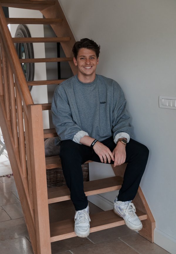 Sudadera gris de gran tamaño, pantalones negros y zapatillas blancas. Individuo sentado en una escalera de madera con acabado natural y suelo de piedra.