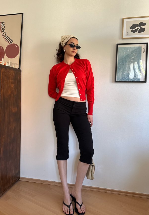Frau trägt eine rote Strickjacke über einem weissen Top, schwarze Caprihosen, schwarze Sandalen, Sonnenbrille und ein beiges Kopftuch und steht drinnen nahe gerahmter Kunst.