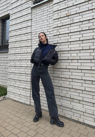 Chaqueta de abrigo negra, suéter de cuello alto azul marino, pantalones vaqueros oscuros de cintura alta y botas negras de tacón grueso. De pie contra una pared de ladrillo blanco texturizado.
