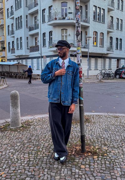Chaqueta de mezclilla sobre una camisa blanca y una corbata de patrón, combinada con pantalones negros holgados y zapatos azul claro. El fondo presenta una escena callejera.
