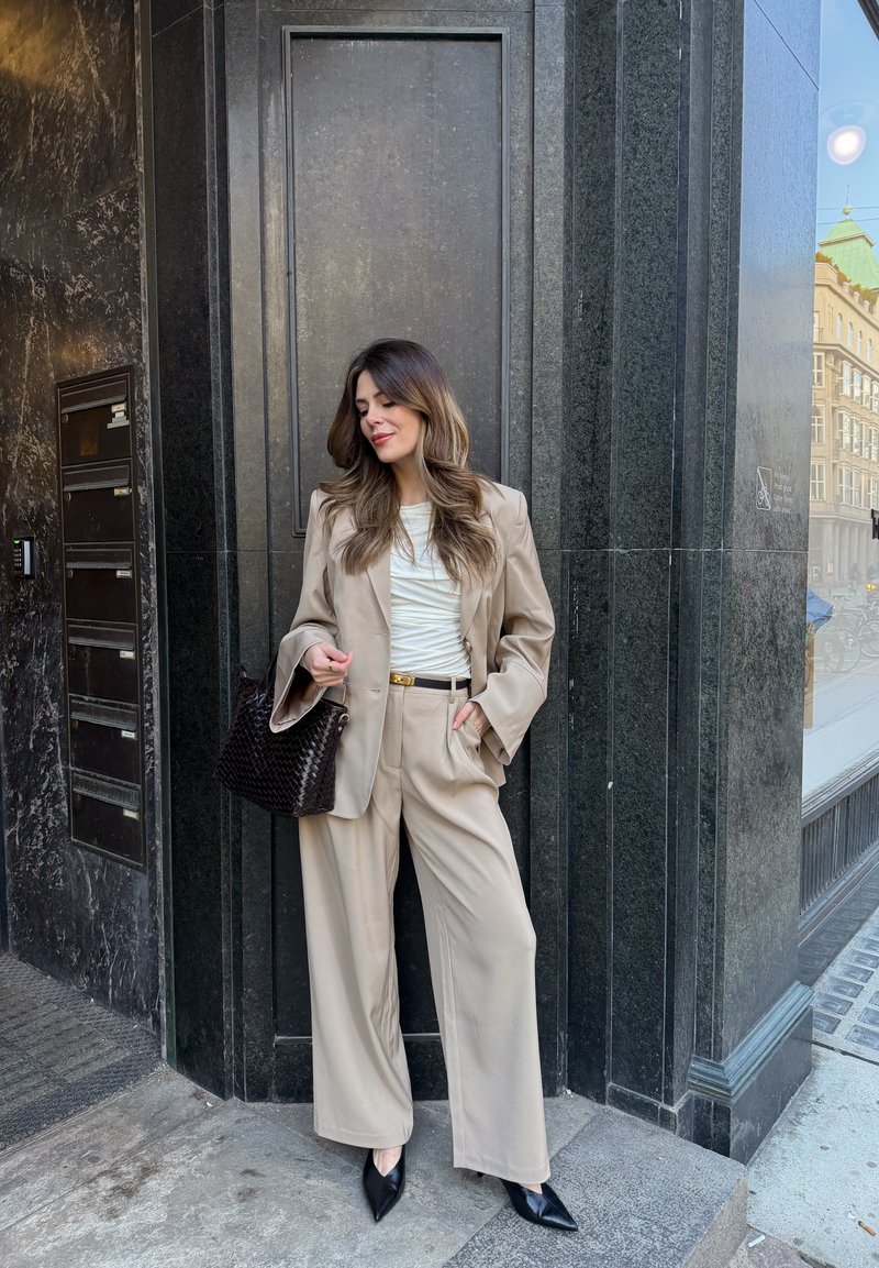 Woman in beige suit and white top stands against dark marble wall, holding black woven handbag, looking down with hand in pocket.