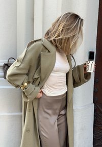 Femme aux cheveux blonds coiffés, portant un manteau beige et un pantalon en satin, tenant un gobelet en papier, debout contre un mur blanc près d'une porte en bois.