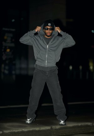 Szürke cipzáros kapucnis pulóver elülső zsebekkel, fekete nadrággal párosítva. Fekete sapkával és napszemüveggel viselve, sötét városi környezetben.