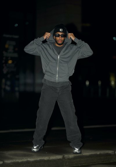 Sudadera con capucha gris con cremallera, pantalones negros, gorro negro y gafas de sol. Zapatillas deportivas con detalles reflectantes; de pie al aire libre por la noche.