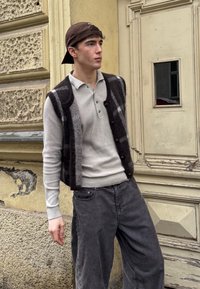 Brown cap, a black and grey patterned waistcoat over a light grey long-sleeve polo shirt, and grey wide-leg trousers, standing against a textured wall.
