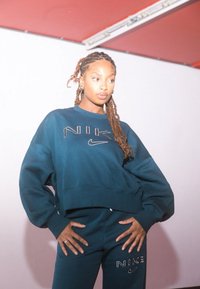 Σκούρο πράσινο cropped sweatshirt της Nike και αντίστοιχες παντελόνες με λογότυπα. Το ύφασμα φαίνεται απαλό, η σχεδίαση είναι χαλαρή με χαμηλωμένους ώμους.