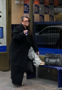 Sort oversized frakke, grå bukser og chunky beige støvler. Holder en kaffekop og står ved siden af en grå håndtaske på en blå bænk.