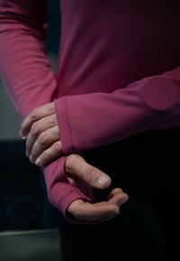 Hands adjusting the thumb hole of a long-sleeve, dark pink sports shirt in a dimly lit environment.