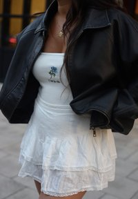 Čierna kožená bunda, biele volánkové šaty s čipkovým lemom a biele tričko s vyšívanými modrými rúžami a textom. Viditeľné spodné vrstvy.