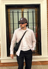 Hemd mit Streifen und langen Ärmeln, weißes T-Shirt darunter, schwarze Hose, braune Mütze mit Logo, dunkle Sonnenbrille, schwarze Tasche über der Schulter, stehend neben einem Fenster.