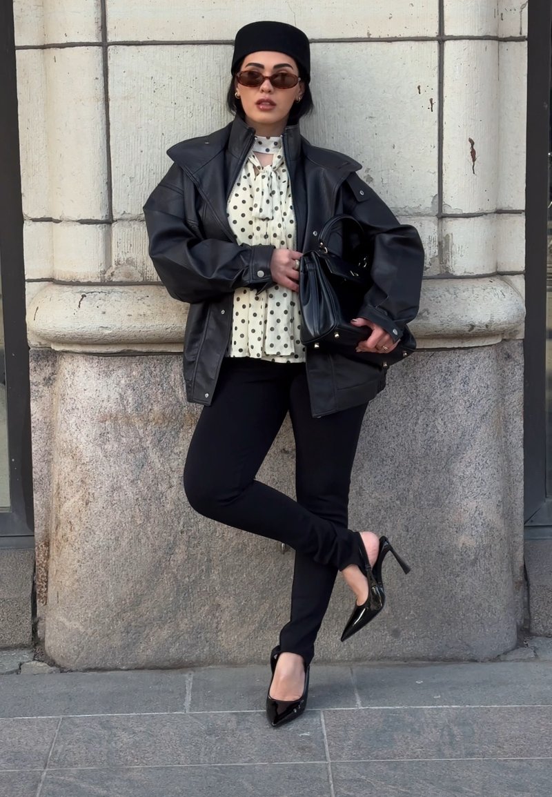 Femme en veste en cuir noire, blouse à pois, pantalon noir, talons, lunettes de soleil et chapeau, appuyée contre un mur en pierre tenant un sac à main noir.