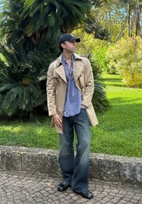 Beige trench coat over a blue and white check shirt, paired with flared dark jeans and black shoes. Green foliage in the background.