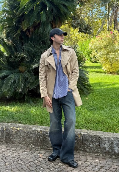 Gabardina beige sobre una camisa de cuadros en azul y blanco, combinada con jeans oscuros de campana y zapatos negros. Follaje verde de fondo.