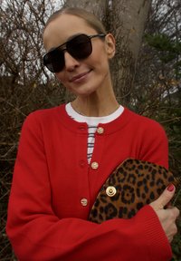 Cardigan rouge avec boutons dorés, chemise rayée en dessous, et pochette imprimé léopard avec un accent doré. Des lunettes de soleil foncées complètent le look.