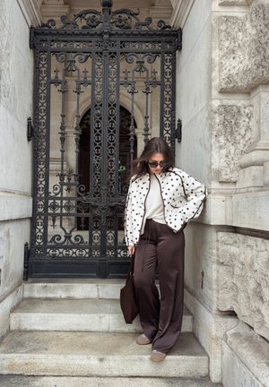 Mujer con chaqueta de lunares y pantalones anchos y oscuros, de pie en escalones de piedra junto a una puerta de hierro negra ornamentada y la pared de un edificio de piedra.