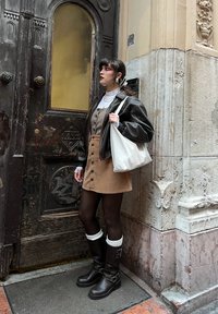 Model iført en brun kordfløjlskjole med knapper, sort læderjakke, sorte knæhøje støvler med detaljer, og holder en hvid totebag.