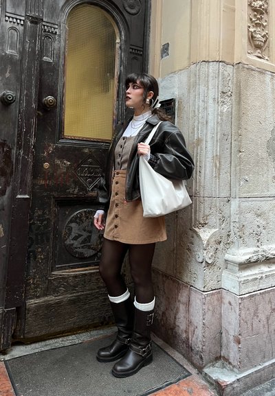Modelo vistiendo un vestido de pana marrón con botones, chaqueta de cuero negra, botas altas negras con detalles y sosteniendo un bolso tote blanco.