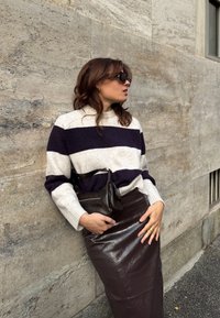Donna che indossa un maglione a righe, una gonna di pelle e occhiali da sole, appoggiata a un muro di pietra con una piccola borsa di pelle in vita.