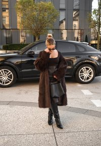 Longue veste en fausse fourrure marron portée sur un haut noir ajusté et une jupe en cuir, assortie à des bottes noires montantes et un sac à main noir.