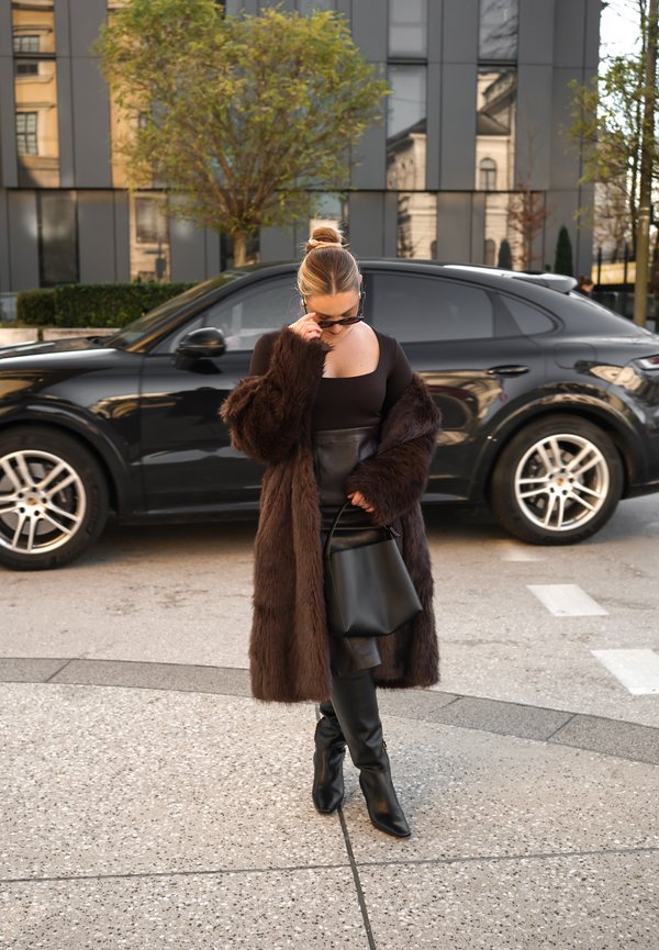 Long manteau en fausse fourrure marron porté sur un haut noir ajusté et une jupe en cuir, assorti à des bottes noires montant jusqu'à la cuisse et un sac à main noir.
