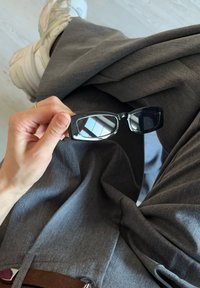 Occhiali da sole rettangolari neri tenuti in mano, con montature lucide e lenti specchiate, poggiati su tessuto grigio e accompagnati da una cintura marrone.