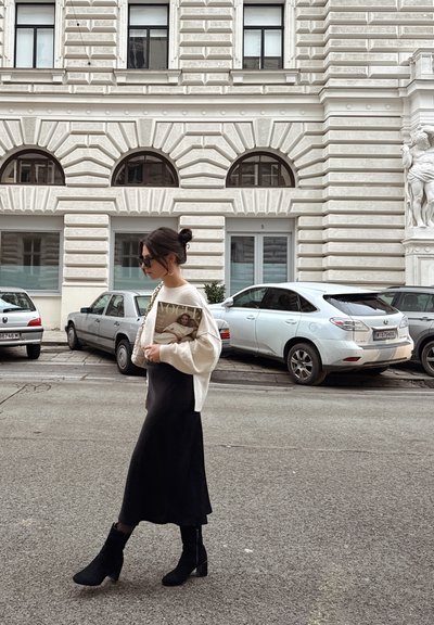 Mujer vestida con un suéter crema y una falda midi negra, sosteniendo una revista, caminando pasado coches y un edificio blanco texturizado con ventanas arqueadas.