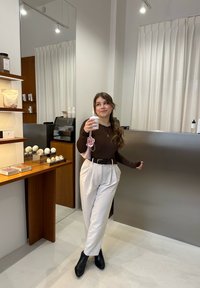 Brauner Langarm-Top, weiße hochtaillierte Hose mit Falten, schwarze Ankle Boots, ein Getränk haltend, in einem modernen Interieur mit einer Holztheke.