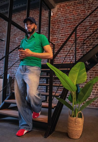 Camisa polo verde, jeans de color azul claro, zapatos rojos. Hombre de pie cerca de unas escaleras, sosteniendo un teléfono. Planta en maceta en una cesta trenzada a su lado.