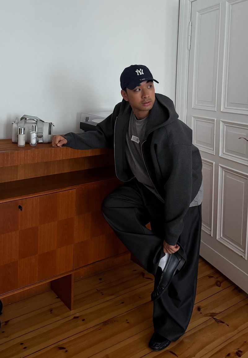 Jeune homme portant un sweat à capuche foncé et une casquette ajuste sa chaussure noire en s'appuyant sur une armoire en bois dans une pièce avec des portes à panneaux blanches.