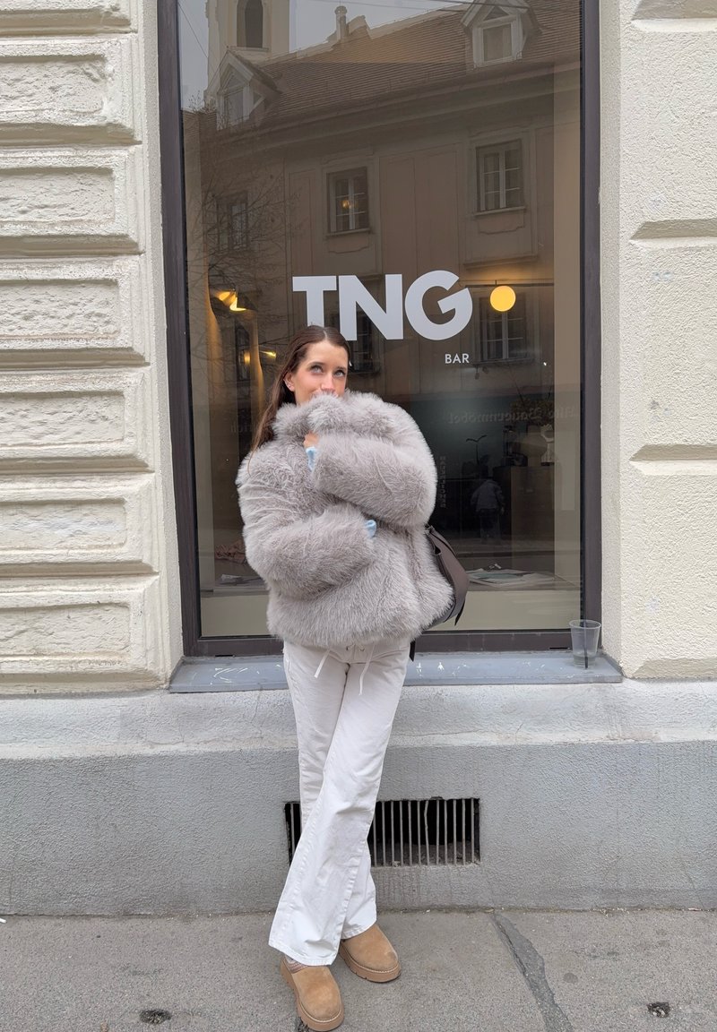 Jonge vrouw in witte broek en grijze bontjas staat buiten de TNG Bar met haar hand bij haar gezicht.