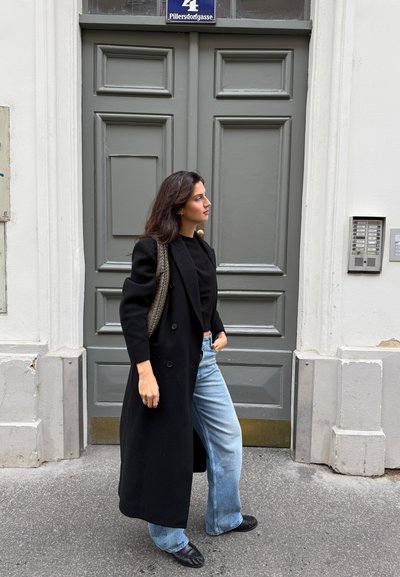 Abrigo negro largo sobre una blusa negra, jeans acampanados de azul claro y zapatos negros. Bolso de mimbre marrón al hombro; de pie frente a una puerta gris.