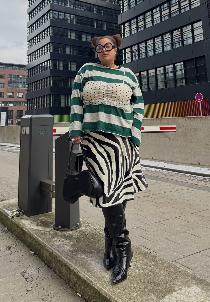 Person with round glasses and two buns wearing a green striped sweater, polka dot top, zebra skirt, black boots, and holding a black bag by buildings.