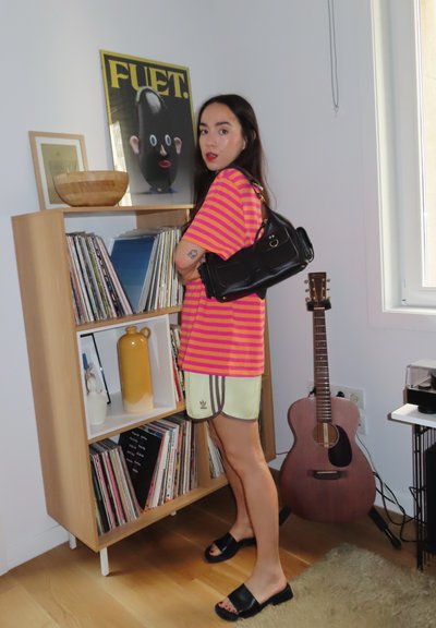 Camiseta a rayas en tonos rosa y naranja combinada con pantalones cortos amarillos claros, chanclas negras y un bolso negro. Muebles y decoración de madera al fondo.