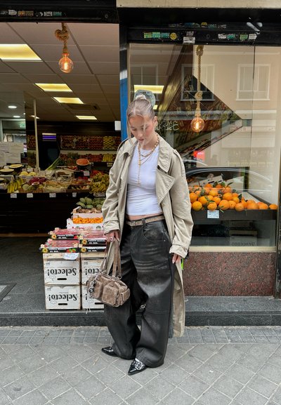 Trench beige sobre una blusa blanca, pantalones anchos negros y un bolso marrón, de pie frente a un mercado con exhibiciones de frutas.