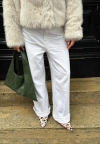 Personne portant un pantalon blanc retroussé, une veste en fourrure crème, des chaussures pointues à pois, et tenant un sac à main en cuir vert foncé, debout sur des marches en pierre.