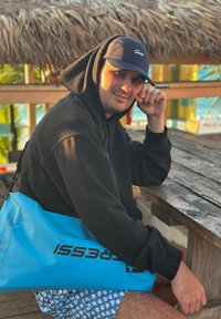 Mann in schwarzer Kapuzenjacke und Kappe sitzt an einem Holztisch im Freien, trägt eine hellblaue Trockenbag, mit Strohdeckendach und tropischem Hintergrund.