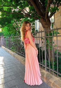 Maxi abito plissettato in rosa pallido con un bustier floreale rosso. Stilizzato con una fessura laterale, indossato con accessori rossi su uno sfondo verde.