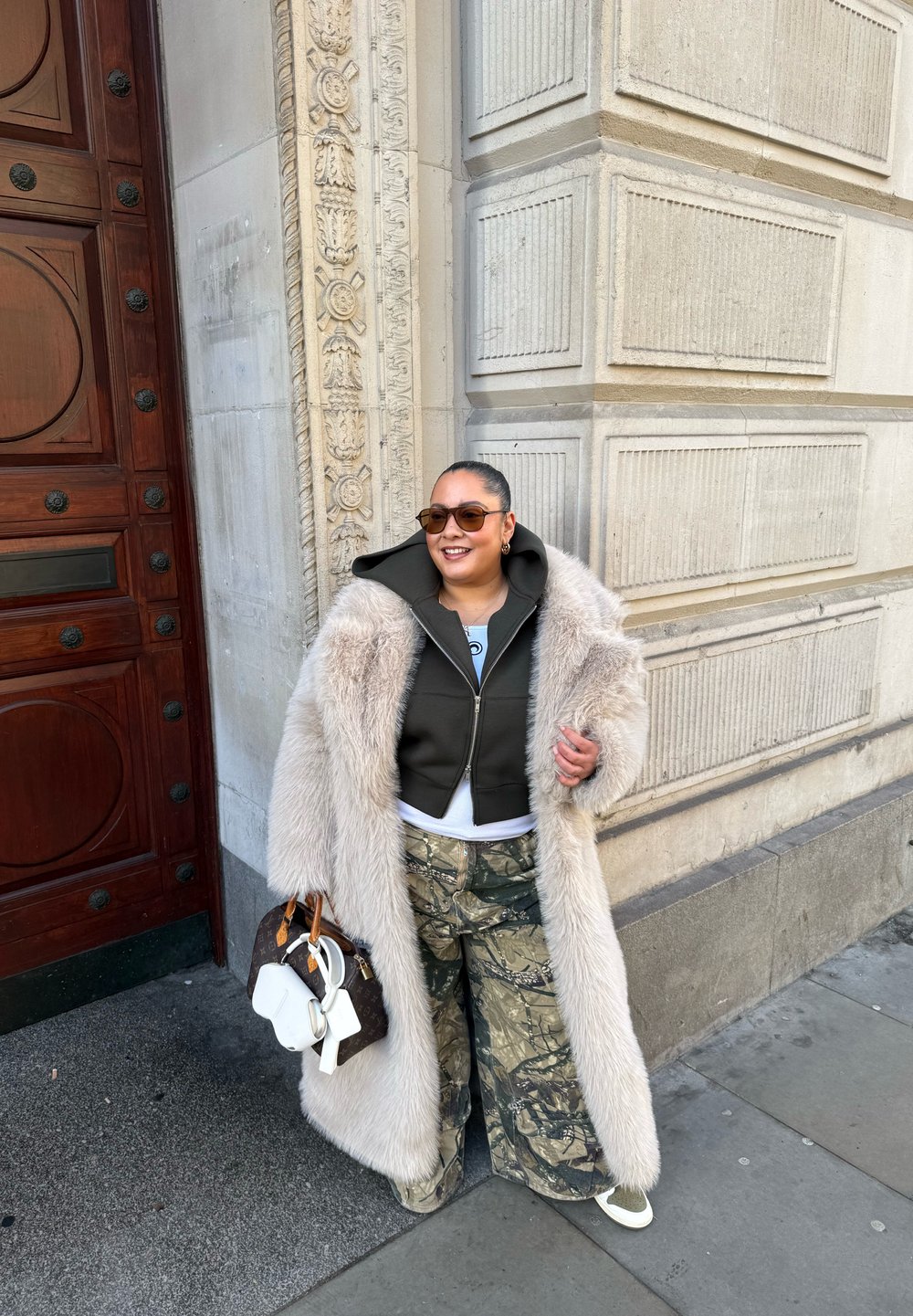 Long manteau en fourrure grise claire porté sur un sweat à capuche court vert foncé, chemise blanche et pantalon large camouflage. Sac à main marron avec des accents blancs.